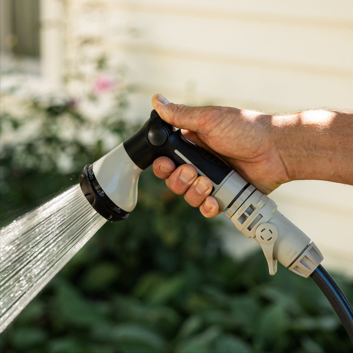 A close-up of a hand holding a garden hose spray nozzle, spraying water outdoors in front of a blurred house and garden background.