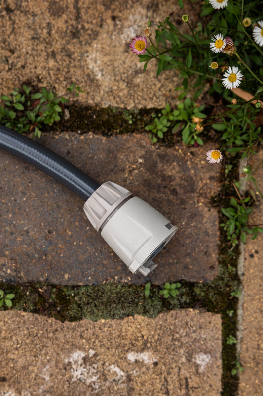 A close-up of a gray Hose End connector on the end of a charcoal hose, placed on a stone paver outdoors with moss, weeds, and small white flowers around it.