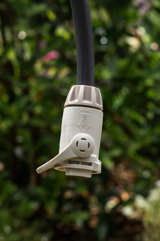 A close-up of a warm grey outdoor garden hose end connector with flow control with a charcoal hose attached, set against a blurred green garden background.