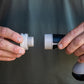 Close-up of a person's hands assembling a grey threaded accessory connector onto a handheld spray nozzle, with a blurred dark green background.
