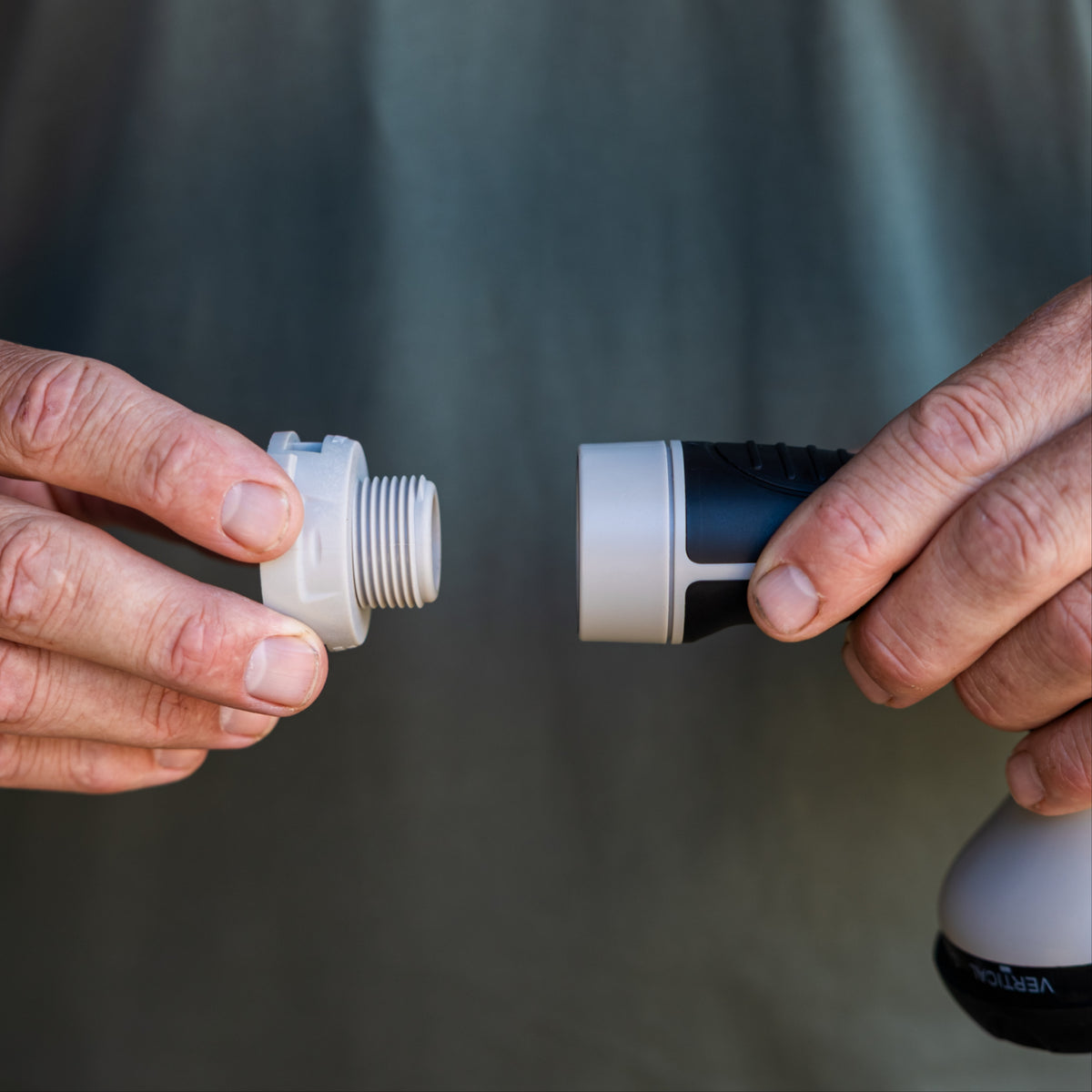 Close-up of a person's hands assembling a grey threaded accessory connector onto a handheld spray nozzle, with a blurred dark green background.