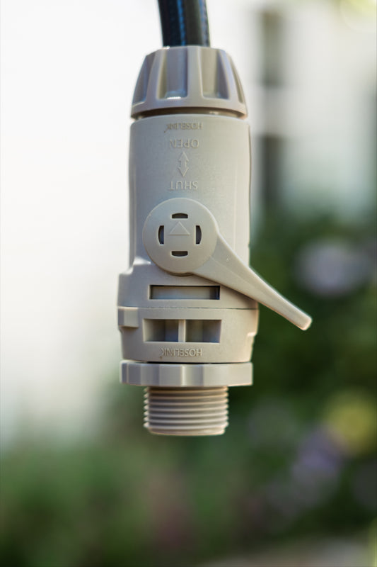 Close-up of a warm gray accessory connetor with swivel and hose end with flowc control, shown outdoors against a blurred green and white background.