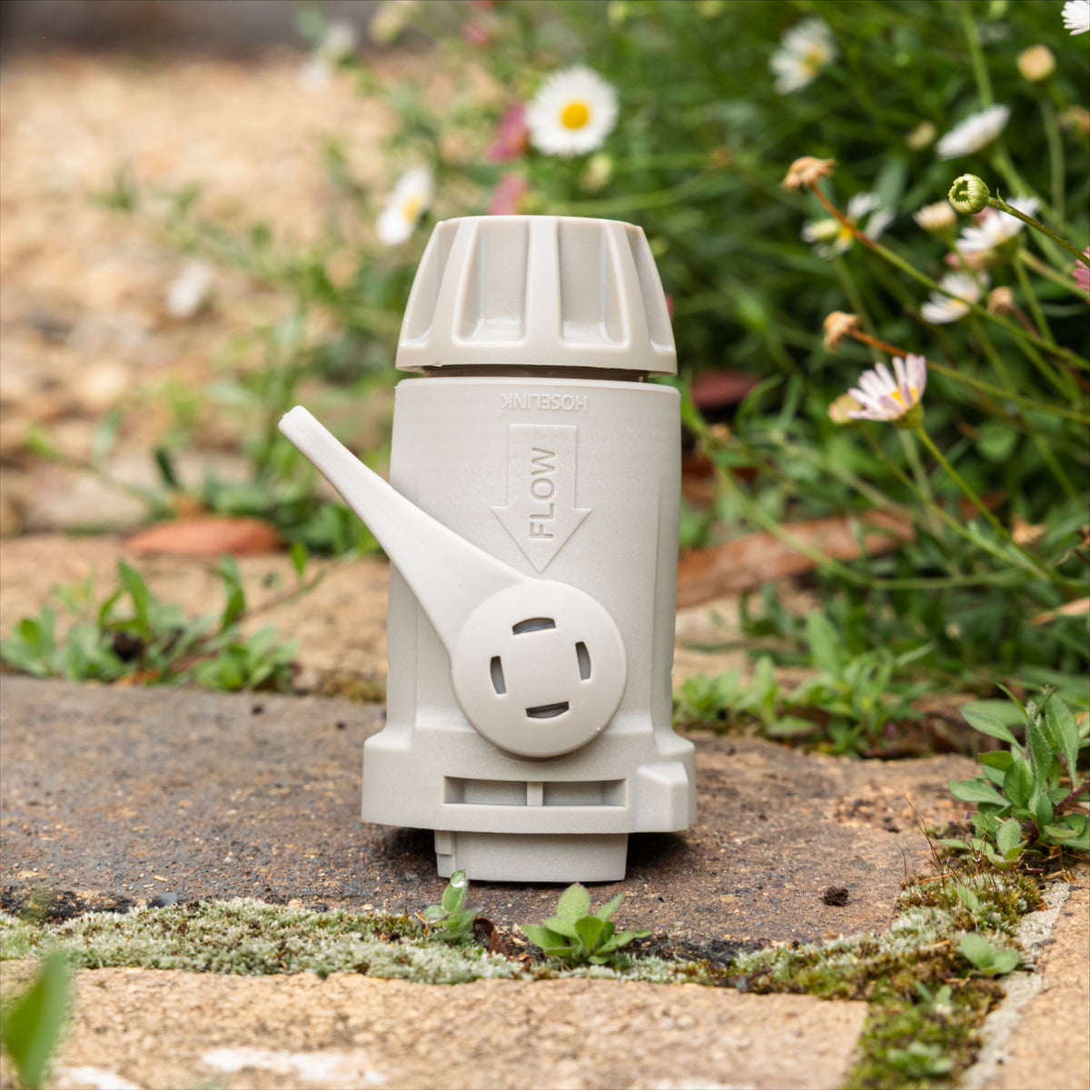 A warm grey plastic garden hose connector weith flow control lever standing on a stone path outdoors with green grass and small white flowers blurred in the background.