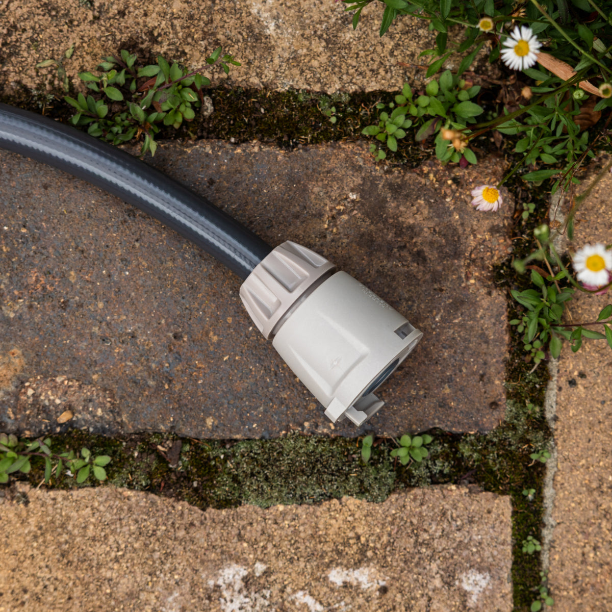 A close-up of a gray Hose End connector on the end of a charcoal hose, placed on a stone paver outdoors with moss, weeds, and small white flowers around it.