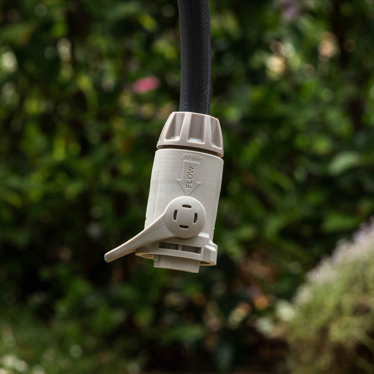 A close-up of a warm grey outdoor garden hose end connector with flow control with a charcoal hose attached, set against a blurred green garden background.
