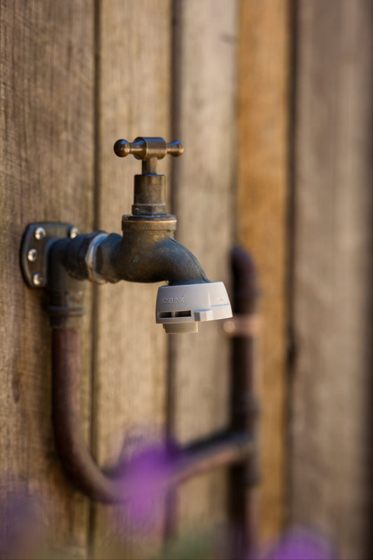 A close-up of an outdoor tap with a grey Hoselink Universal Tap Connector attached to the thread, set against a rustic wooden wall background.