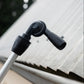 A close-up of a black gutter cleaner head attachment mounted on the silver telescopic cleaning system pole, set against a corrugated metal roof background.