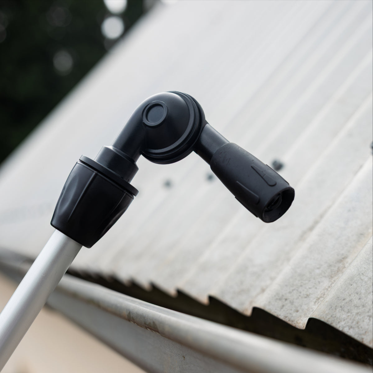 A close-up of a black gutter cleaner head attachment mounted on the silver telescopic cleaning system pole, set against a corrugated metal roof background.