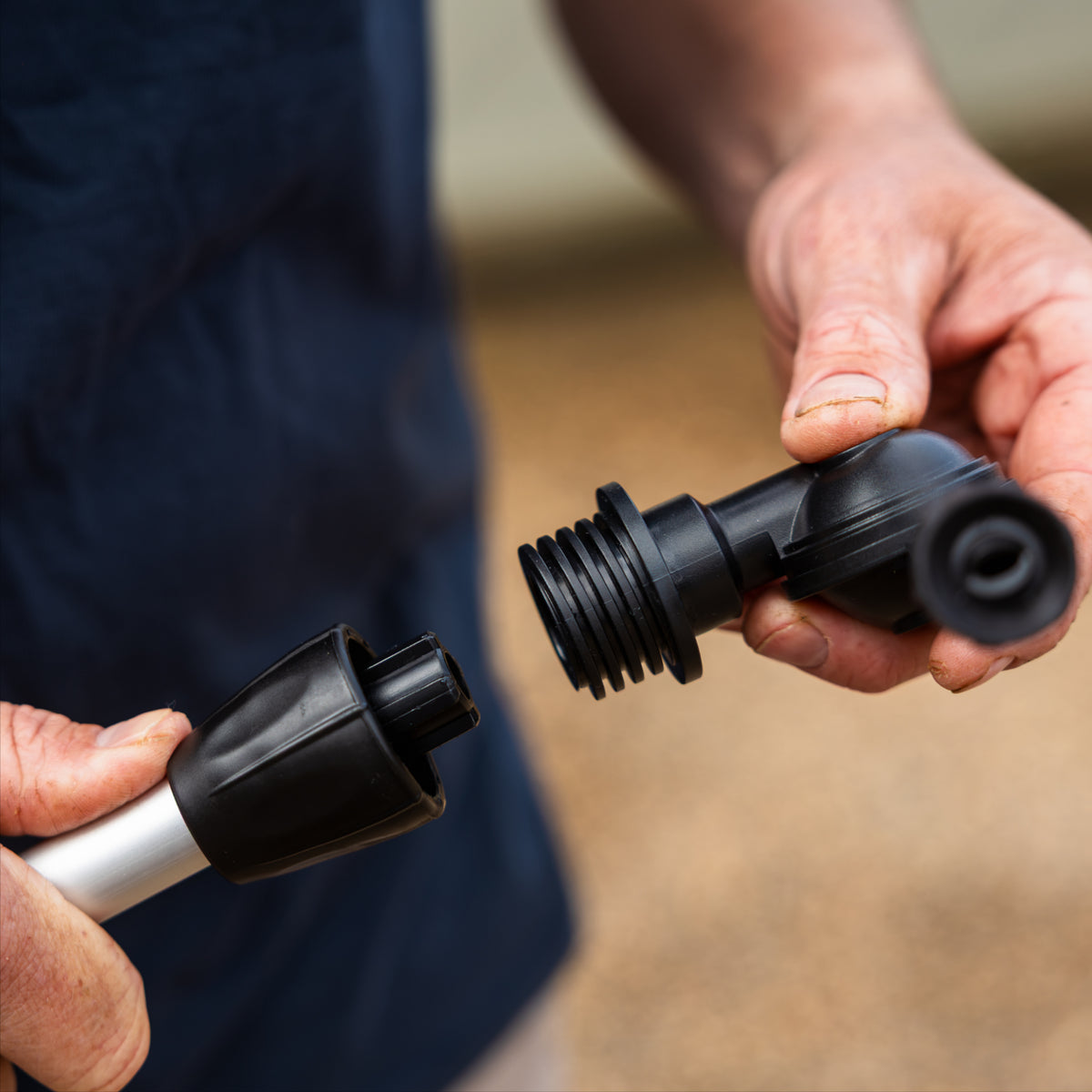A close-up of a person's hands holding  black plastic gutter cleaner head attachment, being installed on the silver metal telescopic claning system poler, against a blurred outdoor background.