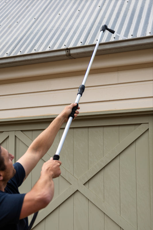 A man in a navy blue t-shirt and khaki pants uses a long-handled gutter cleaner to clean the gutters of a beige garage building with green doors.