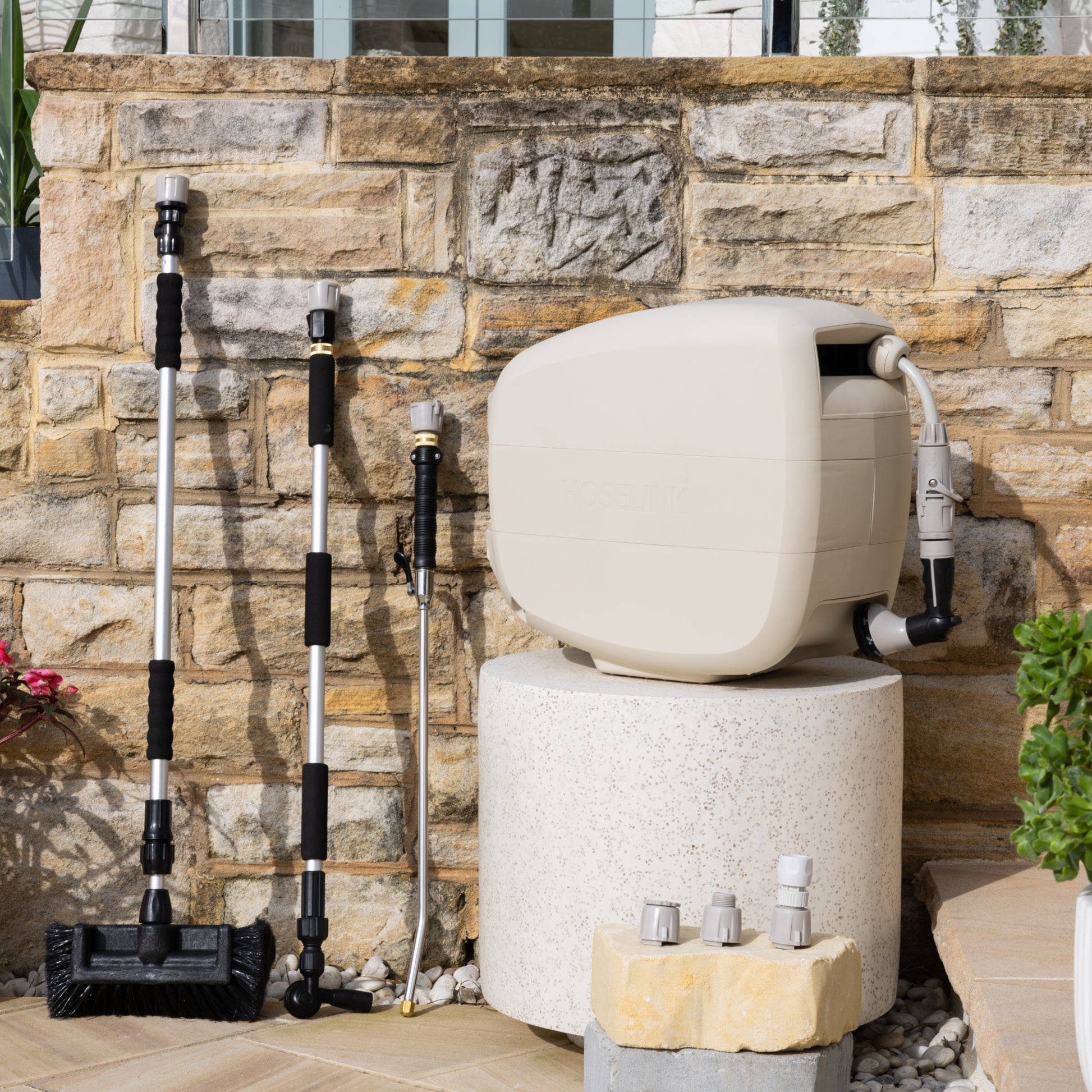 Evolve Beige Retractable Hose Reel on a white stone table with an adapter pack in front and Extendable Cleaning Brush, Super Jet Washer and Pivot Gutter Cleaner leaning on a sandstone wall next to the reel