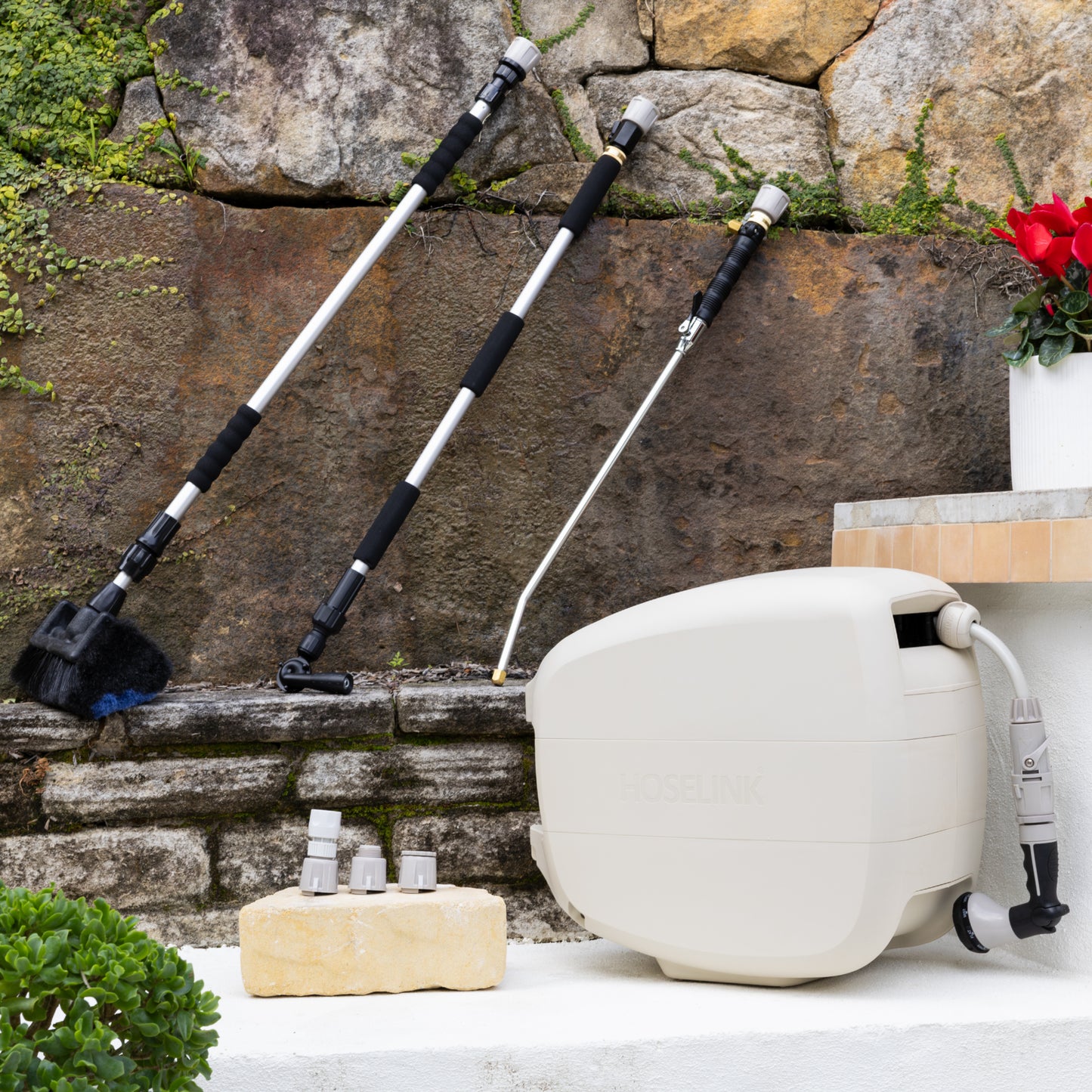 Beige Evolve Retractable Hose Reel sitting on a white step with an Extendable Cleaning Brush, Pivot Gutter Cleaner and Super Jet Washer Leaning against a stone wall and a warm grey Adapter Pack next to the reel