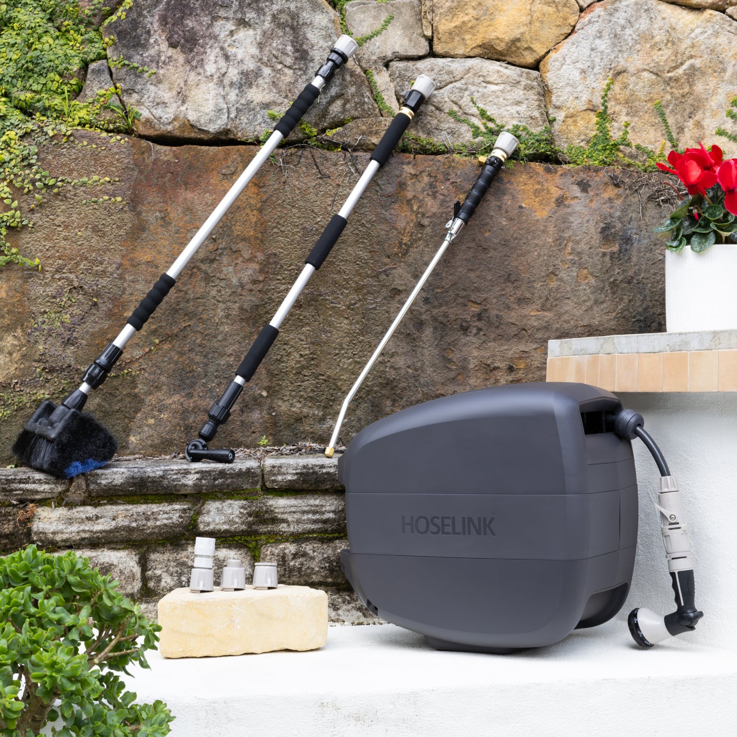 Evolve Charcoal Retractable Hose Reel sitting on a white step with an Adapter Pack next to it and an Extendable Cleaning Brush, Pivot Gutter Cleaner and Super Jet Washer Leaning against a stone wall in the background