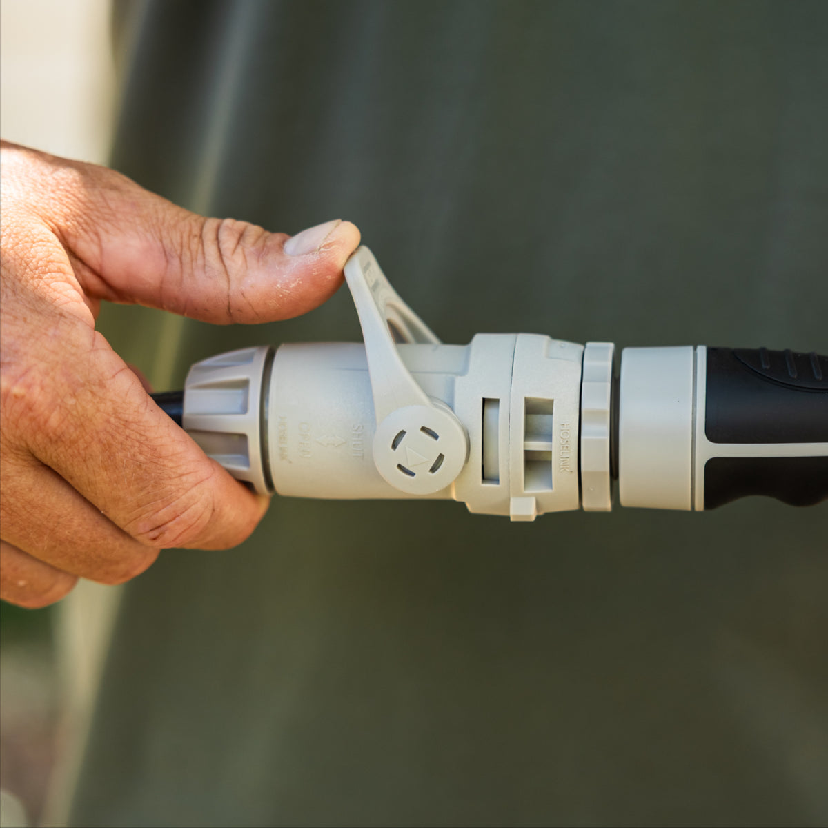 A close-up of a person's hand pressing the flow control lever on a grey Hose end connector attached to a black spray gun