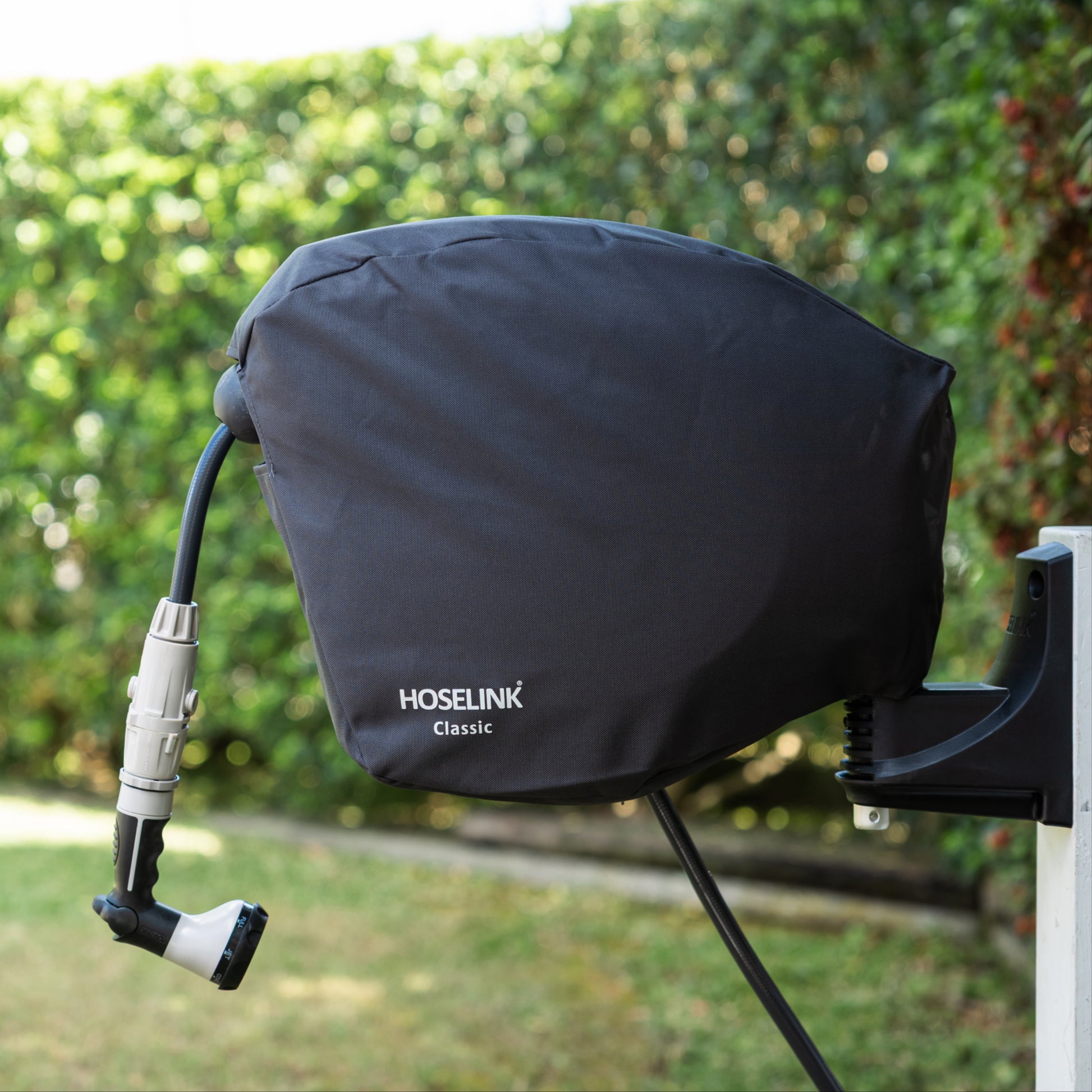 Charcoal HOSELINK Classic outdoor hose reel cover with hose attached to a white post with green lawn and hedges in the background 
