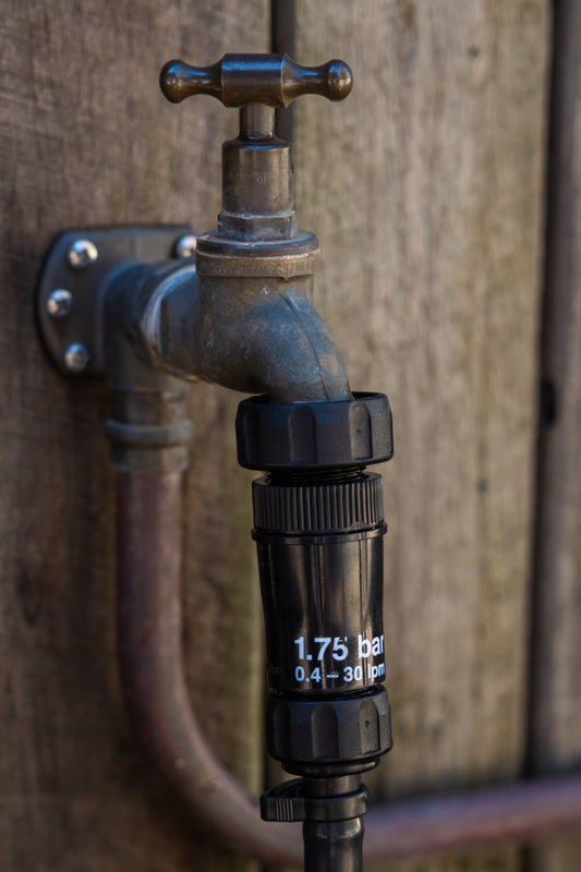 Close-up of black 1" tap adapter connecting irrigation pressure regulator to a brass tap on a wooden fence