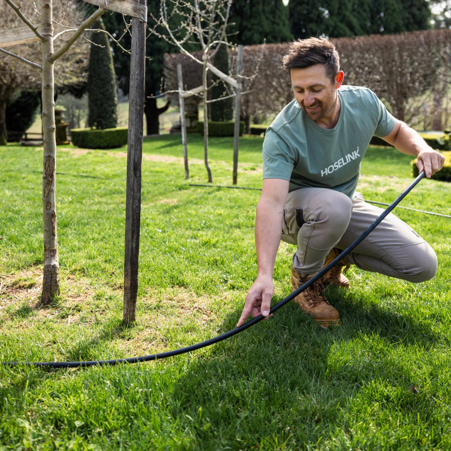 Man in a garden laying out a length of irrigation poly pipe around trees on green lawn