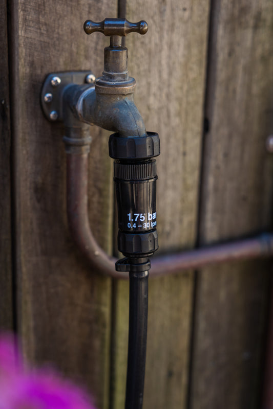 irrigation mit pressure regulator screwed onto a brass tap and connected to black irrigation tubing in front of a wooden fence