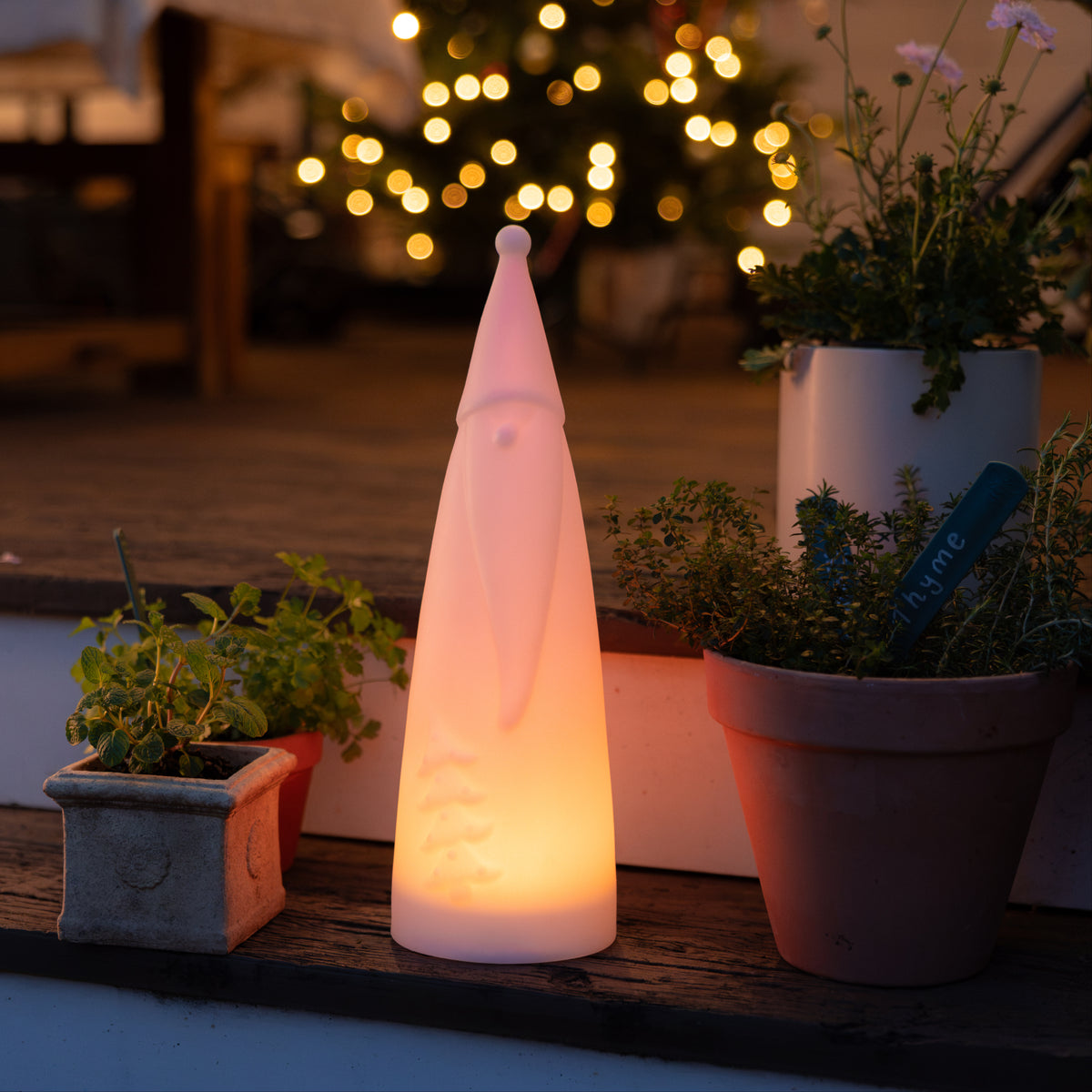 A Tall Solar Santa light, sitting on some wooden stairs. In the middle of 2 pot plants. The Tall Solar Santa light is emitting a peach coloured glow. 