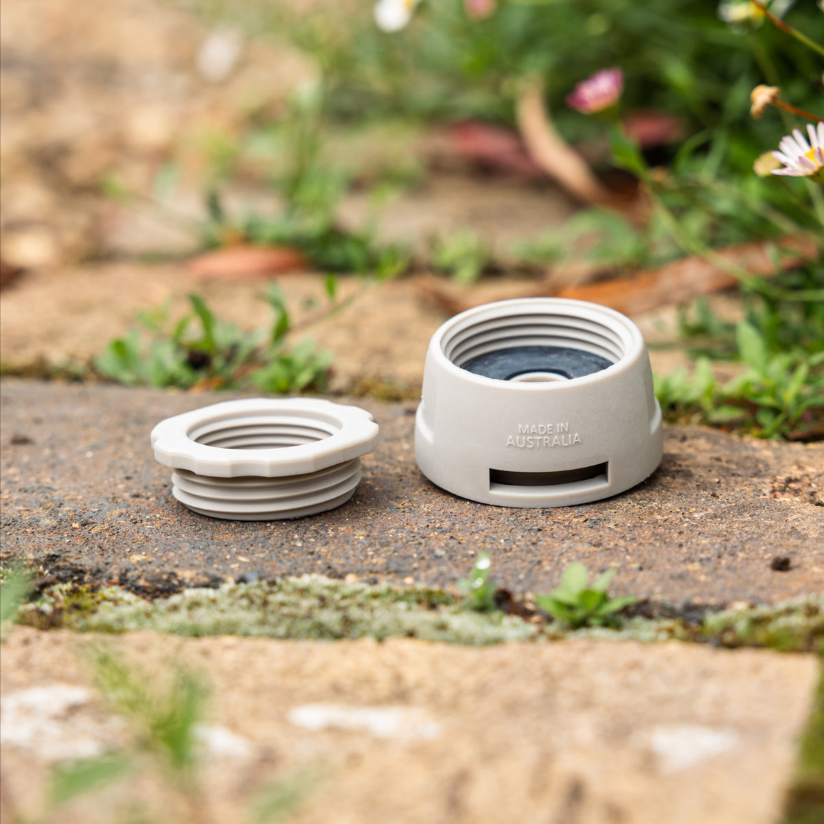 Close up of Gen 2 Universal Tap Connector with reducer removed and placed next to body. Resting on brown paving with blurred flowers in the background