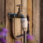 A close-up outdoor tap with a brass handle mounted on a weathered wooden wall, with a warm gray tap connector and hose connector on braided charcoal hose in front.