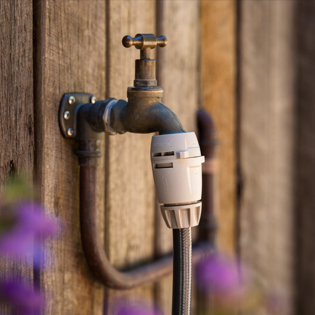 A close-up outdoor tap with a brass handle mounted on a weathered wooden wall, with a warm gray tap connector and hose connector on braided charcoal hose in front.