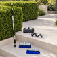 A telescoping window cleaning pole and several brush attachments arranged on outdoor stone steps beside trimmed green hedges. The set includes a long silver-and-black pole with black rubber grips and blue/gray brush heads, plus a black squeegee and two black nozzle attachments.