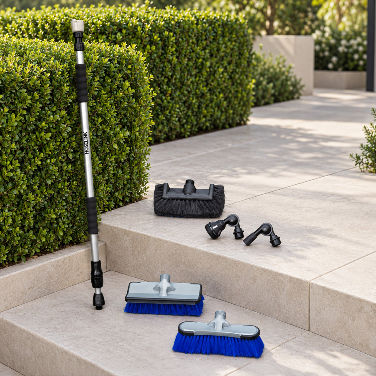 A telescoping window cleaning pole and several brush attachments arranged on outdoor stone steps beside trimmed green hedges. The set includes a long silver-and-black pole with black rubber grips and blue/gray brush heads, plus a black squeegee and two black nozzle attachments.