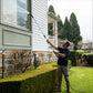 A man in a backyard uses a telescopic cleaning system with 8-pattern sprayer attachment to clean the siding of a house. The house has white horizontal siding, a stone foundation, and tall green trees in the background.