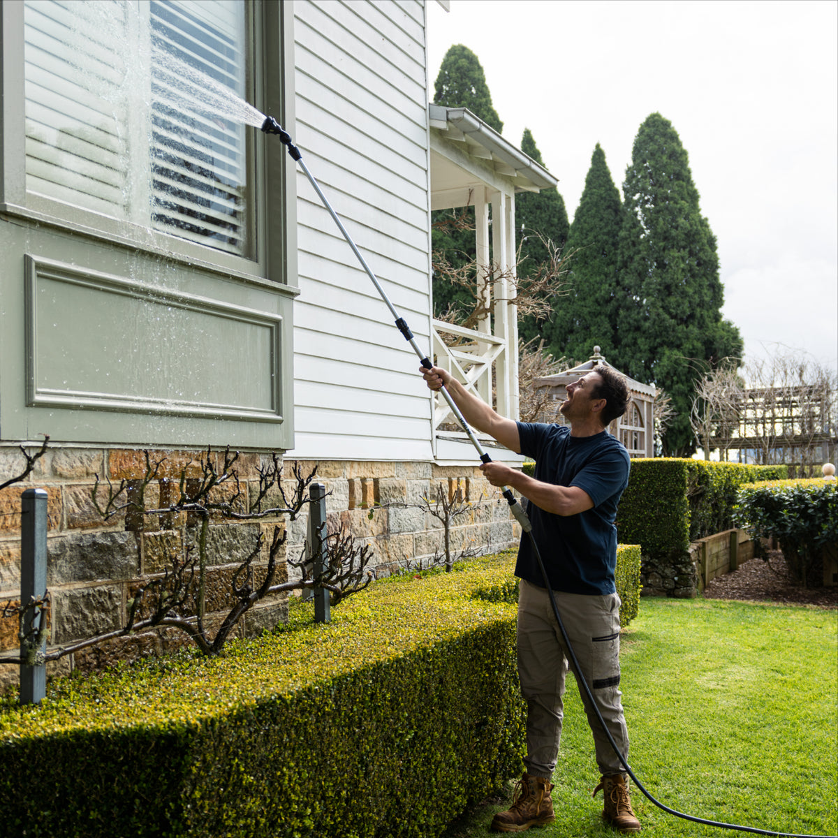 A man in a backyard uses a telescopic cleaning system with 8-pattern sprayer attachment to clean the siding of a house. The house has white horizontal siding, a stone foundation, and tall green trees in the background.