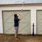 A man in a navy blue t-shirt and khaki pants uses a long-handled gutter cleaner to clean the gutters of a beige garage building with green doors.