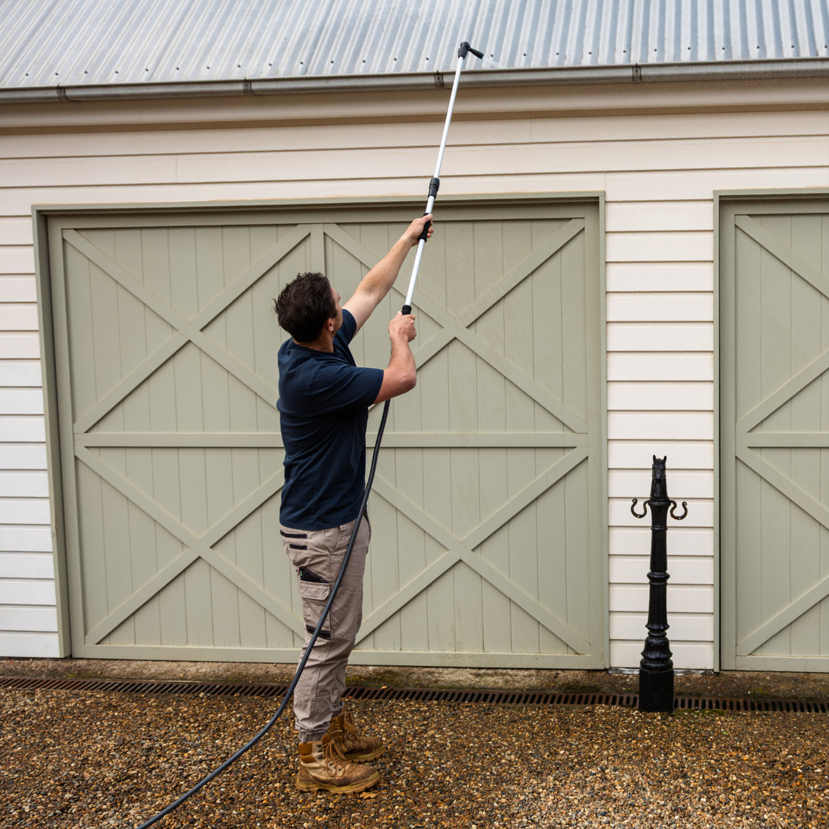 A man in a navy blue t-shirt and khaki pants uses a long-handled gutter cleaner to clean the gutters of a beige garage building with green doors.