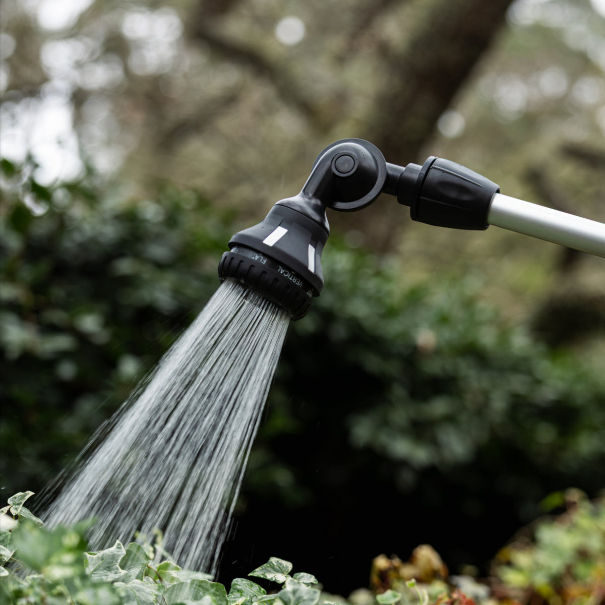 A black adjustable garden hose sprayer with a flexible head spraying water, attached to a silver pole in an outdoor garden setting with blurred green foliage.