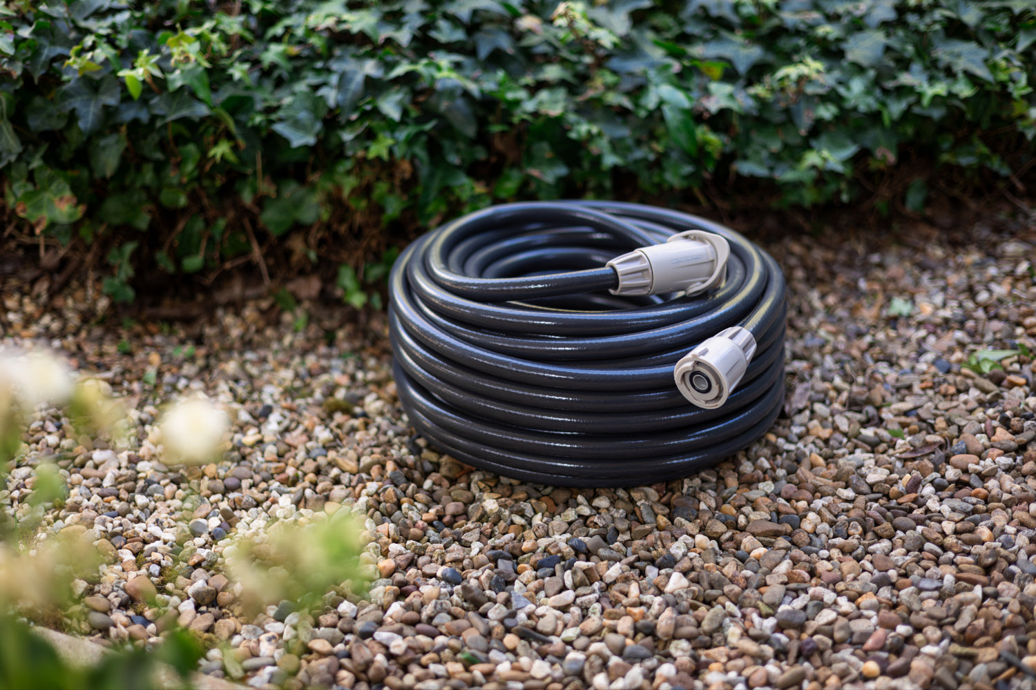 Coiled charcoal coloured garden hose on a pebbled surface with greenery in the background