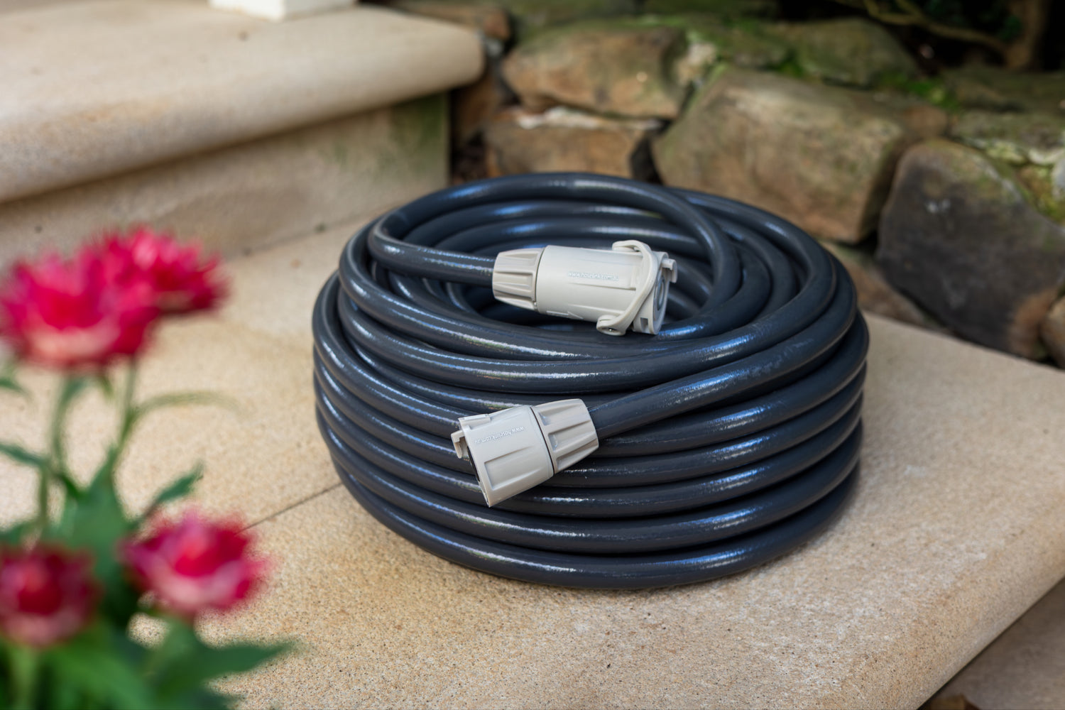 Coiled black garden hose with gray connectors on a stone surface next to pink flowers.