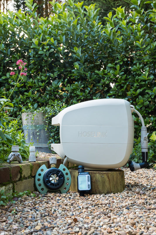 Beige evolve hose reel, 9-pattern sprinkler, automatc tap timer, easy turn 2-way adapter and click-on adapter set sitting on a low brick garden wall surrounded by gravel with a green hedge in the background