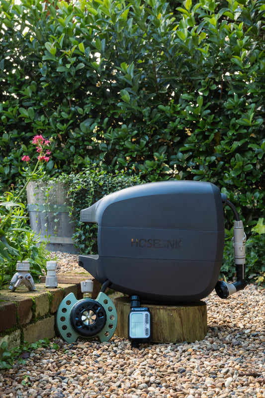 Charcoal evolve hose reel, 9-pattern sprinkler, automatc tap timer, easy turn 2-way adapter and click-on adapter set sitting on a low brick garden wall surrounded by gravel with a green hedge in the background